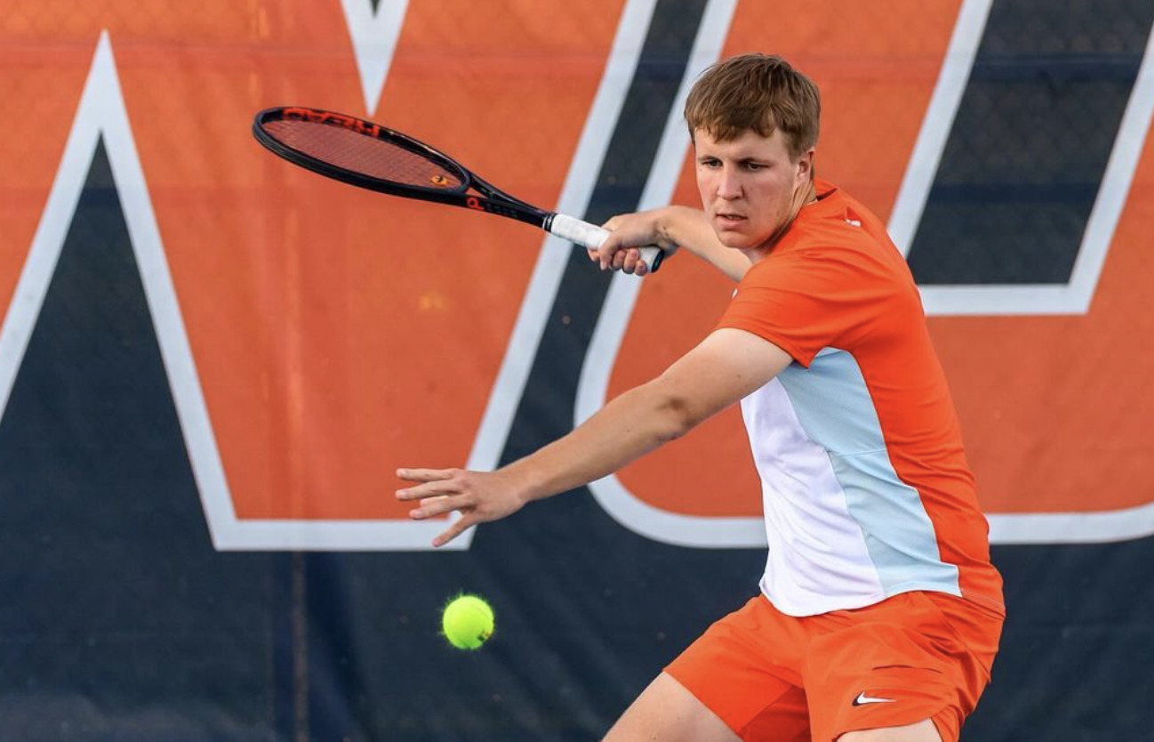 University of Illinois men's tennis player Karlis Ozolins, a tall white man, wears orange and white athletic clothes. He is about to his a tennis ball with his racquet; he is staring at the ball.